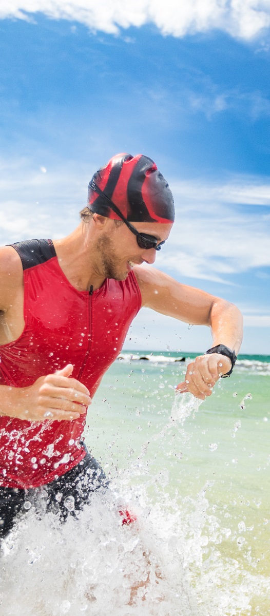 Smartwatch triathlon swimming sport man finishing swim checking heart rate on smart watch. Male triathlete swimmer running out of ocean. Professional athlete in triathlon suit training for ironman.